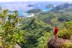 Young female traveller standing at the edge of the cliff at Morne Blanc View Point, overlooking Mahe Island coastline with lush tropical vegetation and crystal blue ocean, Seychell